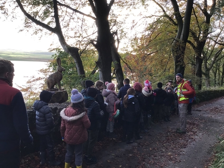 Group of pupils listening to the sounds of nature under trees with lake to one side of them
